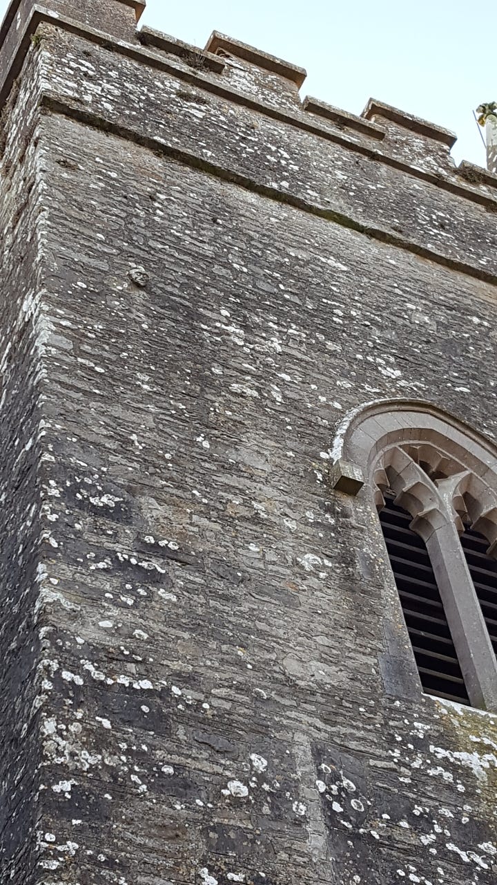 1. old font and standing stone in the graveyard. 2. The medieval tower. 3. Close-up of arched window high on tower wall with carved figure slightly above and to the left. 4. Zoom capture of the stone head with very weathered body barely visible.
