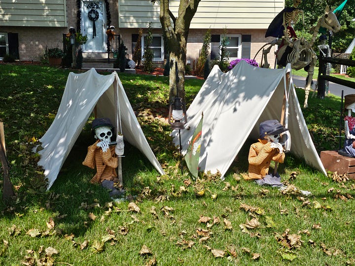 Halloween decorations, including skeletons sitting outside a tent, a skeleton in a coffin, and skeletons attending a funeral.