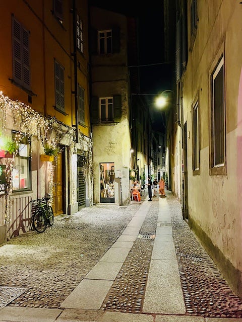 piazza full of tables and people, cobblestone streets, Via Vitani, yellow Italian stucco buildings with green doors, white Christmas lights in summer.