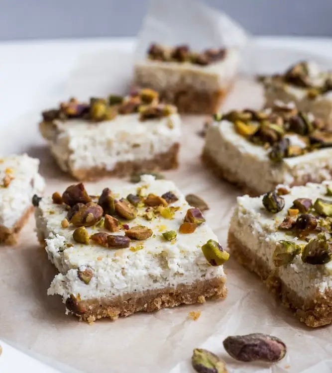 Cardamon & Pistachio Cheesecake Bars cut in squares sitting on parchment and big Rasmalai cake covered in thick whipped cream covered in ground pistachios