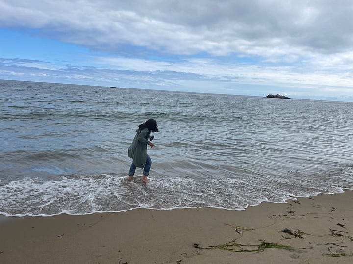 Two photos taken seconds apart. Jen is in jeans and a green sweatshirt-jacket. In one photo she is standing in the shallow white surf to her ankles only. In the next, she lifts her hands over her head as she tries to avoid the small shift in the water that had reached the bottom of her jeans.