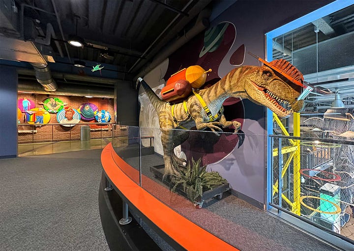 Large dinosaur model wearing a jetpack and a wide pathway through a children's exhibit.