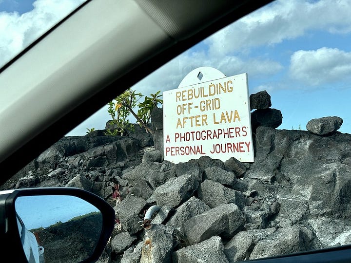 Hand-painted signs placed on lava rock reading "Hot Foot Photography: Current Updates, Photo Video Area Info" and "Rebuilding Off-Grid After Lava: A Photographer's Personal Journey"