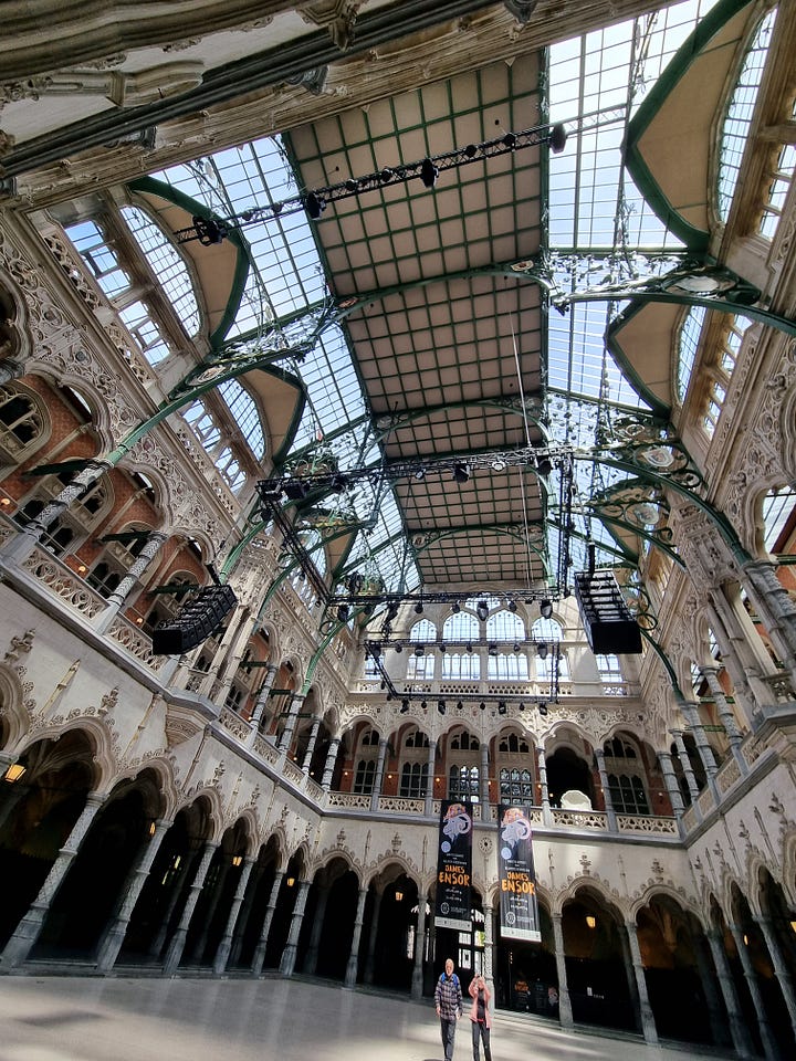 Photo 1: Fort Het Seen in the background with a statue of Lange Wrapper (a water spirit) towering over humans in the foreground. Photo 2: Antwerp Town Hall and statue of Brabo in the foreground throwing a severed hand. Photo 3: Photo from inside the Antwerp Bourse. Photo 4: Dave going down the wooden escalator in the St Anna tunnel. 