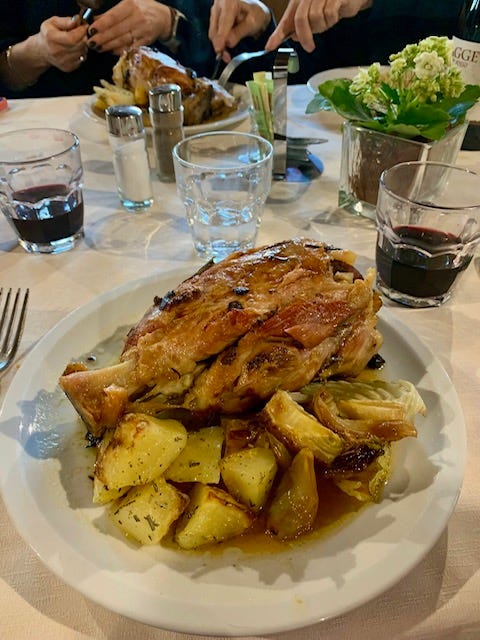 people of all agres sitting around a table in italy, drinking wine, smiling, talking, eating pork shank and potatos