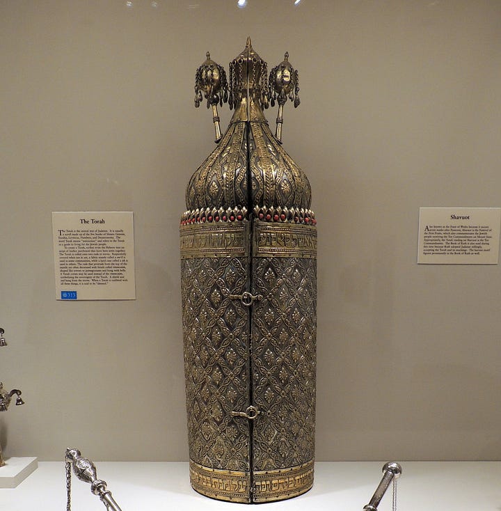Left: Sephardic Torah in elaborate metal case; right: Ashkenazi Torah in maroon mantle with gold embroidery and fringe