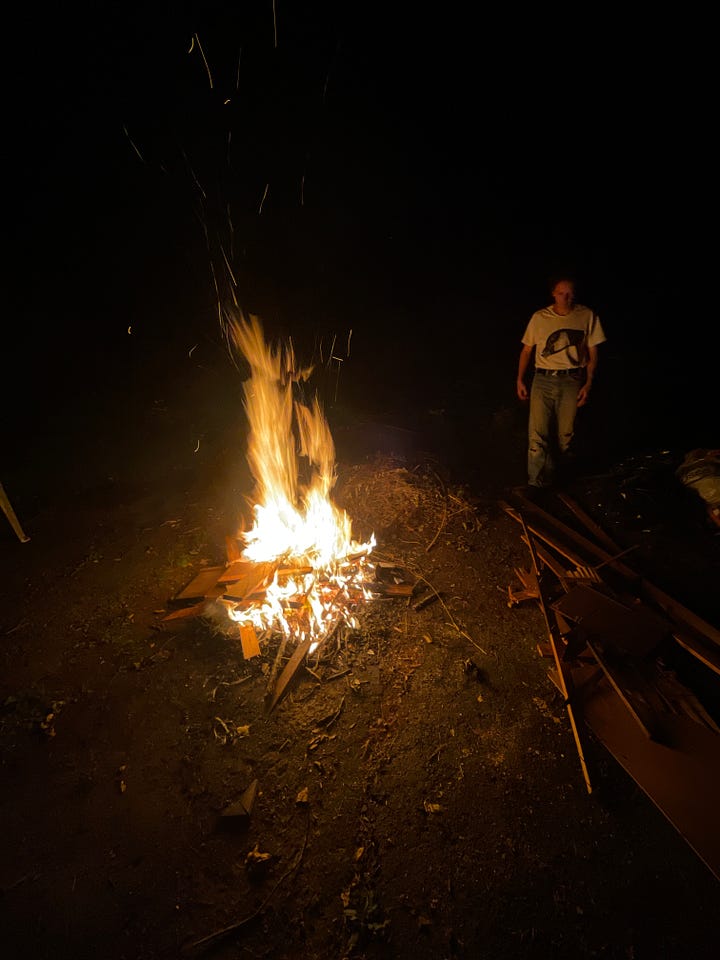 A dog house burning on a bonfire at night, and Liam standing next to a bonfire.