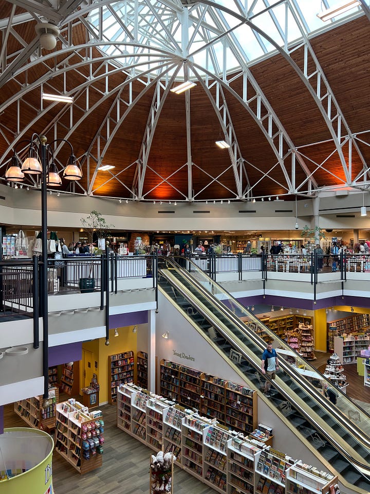 Top left: A wide shot of a large bookstore with a glass ceiling. Top right: A shot of a set of full bookshelves at the bookstore. Botton left: A wide shot of the bookstore showing the escalator down to the kids' section. Bottom right: A stack of books I purchased, which includes The Yellow Wall-Paper, An Ordinary Violence, The Dog of the North, I Love You So Much It's Killing Us Both, and Big Time.