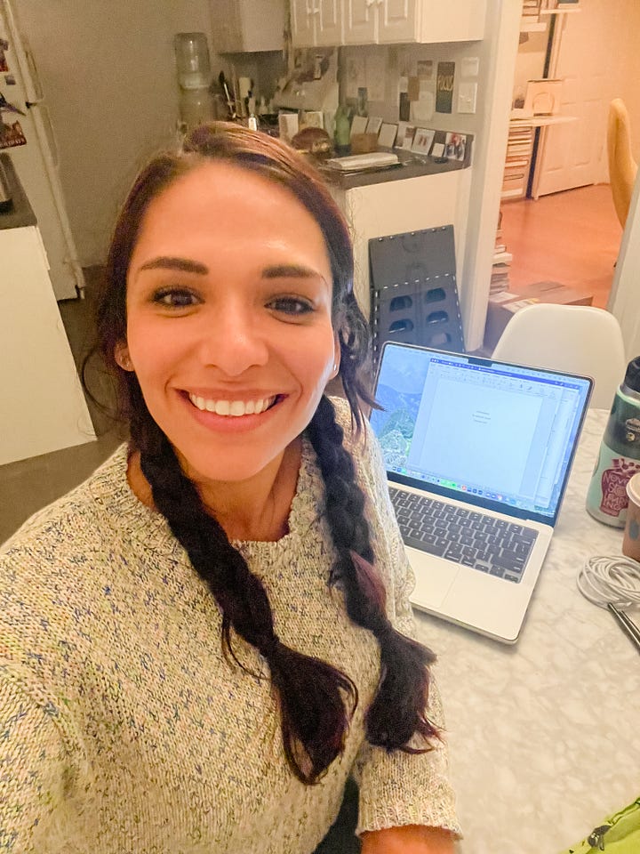 Selfie of woman with laptop behind her, sitting at a kitchen table; mirror selfie photo of woman giving a peace sign with her fingers.