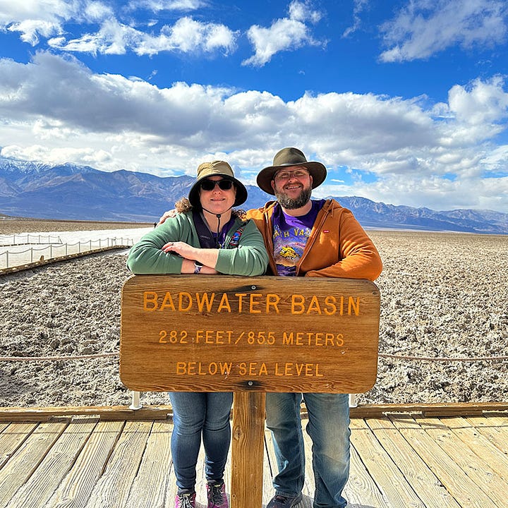 The artist and her husband posing behind the Badwater Basin sign (282 feet below sea level), with a vast salt flat and purple mountains in the background, against a cloud-filled blue sky. The artist and her husband bundled up, with a large snowy volcanic crater in the background. They are smiling but clearly freezing.