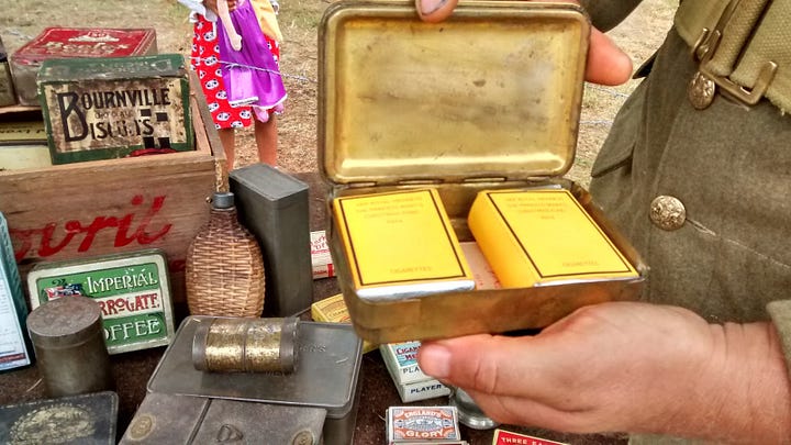 Man in WWI uniform holding open tin to reveal pack of tobacco and pack of cigarettes, men lined up with rifles in WWI uniform with modern kids watching