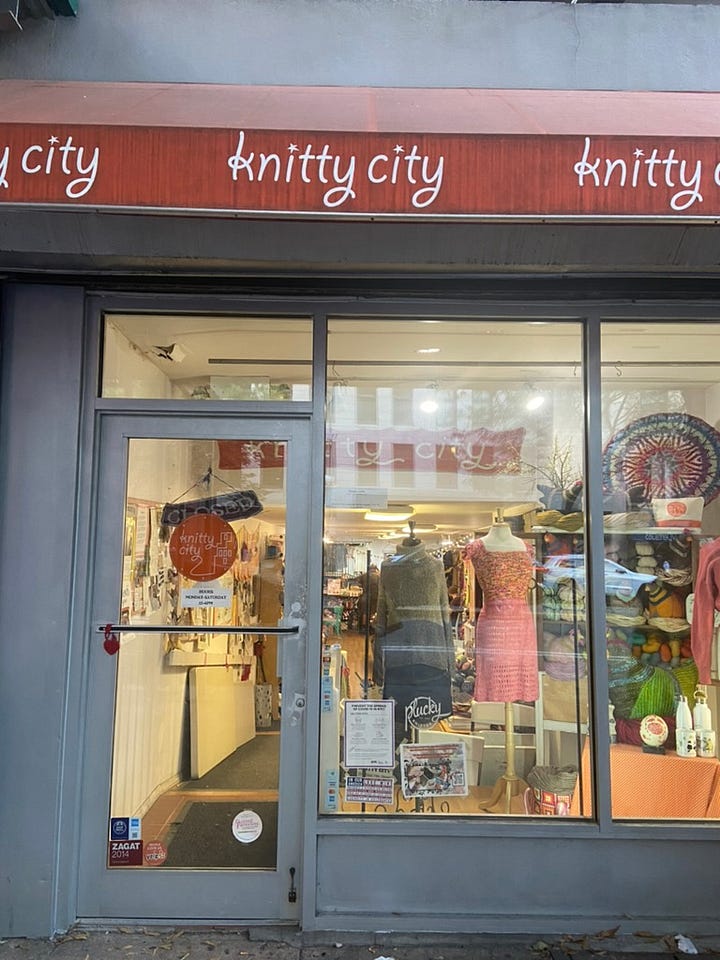 A storefront with a orange sign that says "Knitty City" and some knit garments in the window, a tree trunk covered in some neon green knit.