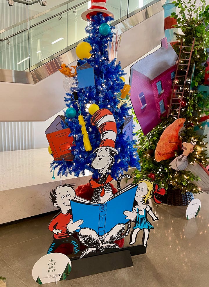 Christmas trees decorated with book characters and elements. On the left, a Christmas tree decorated with characters from Dr. Seuss's Cat in the Hat. On the right, a Christmas tree with a red fire hydrant nestled against it on the left. Decorations on the tree, in reds and yellows, include a red fireman's hat and yellow and orange ribbons intended to look like flames. The book is Fahrenheit 451 by Ray Bradbury. 