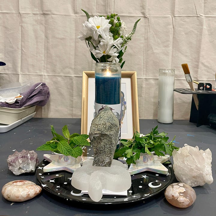 Images are of a simple magical ritual to Aesclepius, involving a green, glass-encased 7-day candle, an icon of Aesclepius healing honey, herbaceous offerings, and healing gems of various sizes, including a snake carved from moonstone with a serpentine point resting on its back.