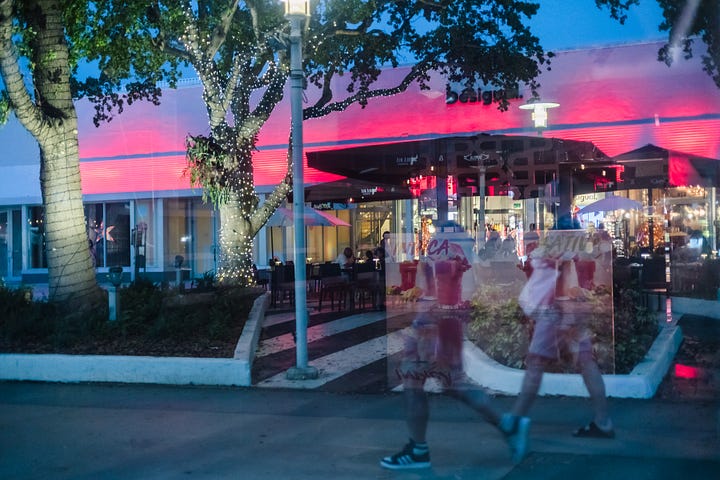 Image of a reflective surface with a round display case for art in a store; photo of a reflective surface overlaid on an outdoor dining and patio scene in a shopping area, with two people walking and superimposed on the background.