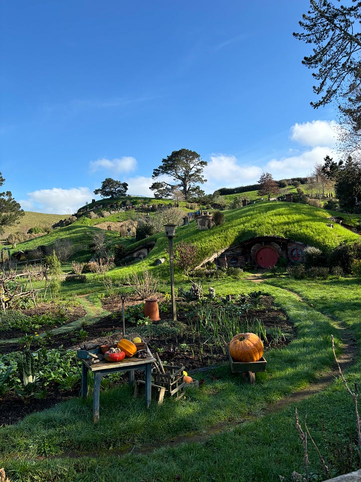 A large garden at the base of Hobbiton with round Hobbit doors tucked into a hillside. A selfie photo of Eija Sumner in front of a blurred background with a green Hobbit door. 