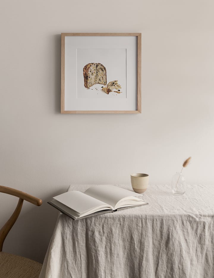 The image on the left shows a framed print of the panettone bread, hanging above a linen covered table with an open book on it, a cup, and a chair to the left. The right image is the original painting on a wood surface with a hand reaching in with a paintbrush. 