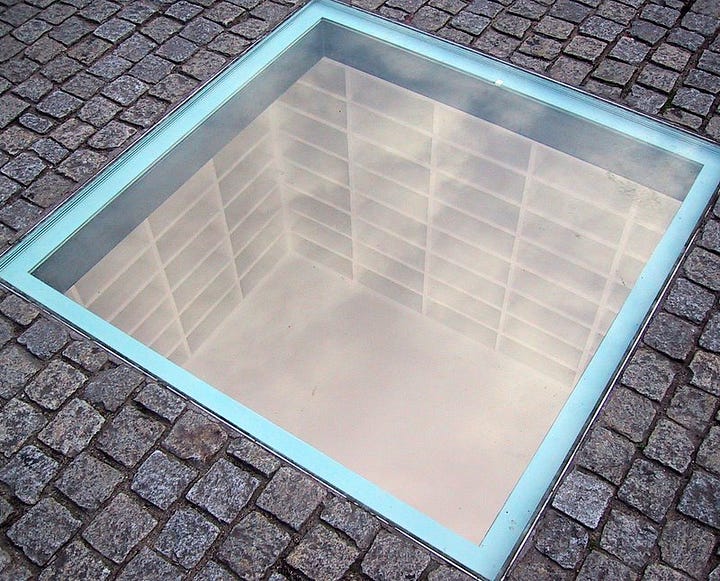 cobbled ground reveals a lit room under the square with empty white bookshelves (left) People's reflections appear in the transparent panel as they look down into the same memorial of empty bookshelves below the surface.