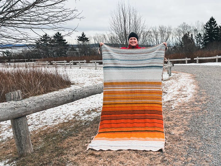 The first image shows 11 balls of yarn as a gradient from chocolate brown through orange, yellow, taupe, and grey, to blues, ending in navy blue, along a store floor. The second photo is the author in black toque, holding a finished crocheted blanket that used the yarn to record the daily high temperatures in 2023. 