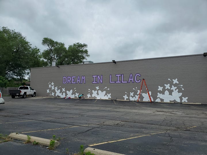 A mural with the words "Dream In Lilac" being painted; a human earthling by a bike