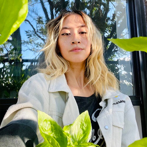 A dark-skinned Black man with a beard and blonde hair stands wearing a bright-blue worker jacket in front of a garden. A thin Taiwanese femme with bleach blonde hair and black wing eyeliner sits on their fire escape surrounded by basil leaves in their garden.