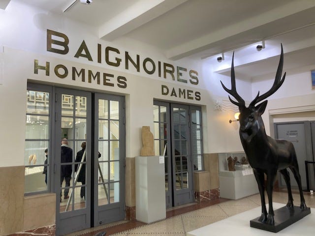 Wide shot of swimming pool gallery; Hommes Dames pool entrance; white marble sculpture of bather; plaster of Paris sculpture of torso with red mushroom cloud in place of the head