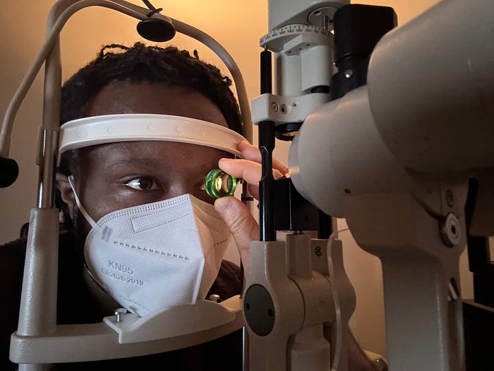 (Image 1) A photo of ArmaniXR's IRL avatar being examined with a slit lamp, which is a beige machine. ArmaniXR holds a circular green-tinted lens up to one eye. He is wearing a white KN95 mask. (Image 2) A close-up photo of ArmaniXR's eye. A reflection of the room can be seen in his iris and pupil. There is a glowing orange light reflected onto his eye.