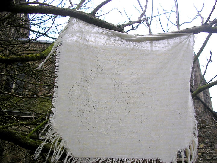 A further away shot of the close up image above of some cream fabric which has pierced holes in it that form an image you can faintly see is a baby holding a sippy cup. You can also see faint writing of the fabric which had a ghostly quality and the fabric has frayed edges and is hung from the branch of an old tree. The next image to the right shows a series of these fabric pieces hanging from the branches of an old tree and flapping in the wind.