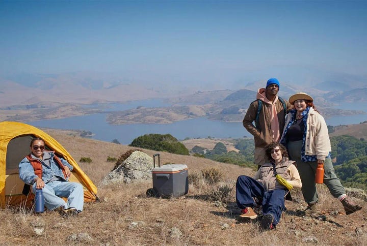 Two photos of several people at a camp site overlooking a lake. In the first photo, there's a cooler in the center of the photo, the sky is hazy blue, and the person on the left is near the edge of the photo. In the second photo, the cooler is gone, the sky is a beautiful sunset, and the person on the left has been moved to the center of the image.