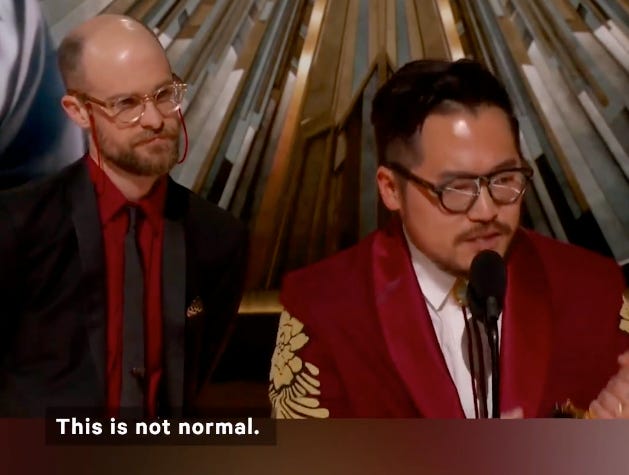 Daniel Kwan at the microphone holding an Oscar with Daniel Scheinert at his side watching