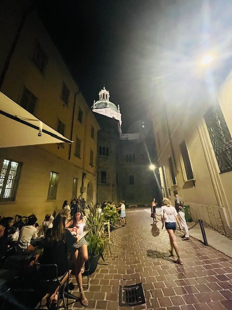 piazza full of tables and people, cobblestone streets, Via Vitani, yellow Italian stucco buildings with green doors, white Christmas lights in summer.