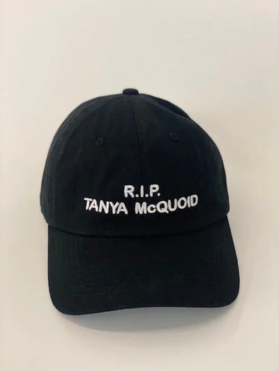 Two black hats with white embroidery. One says, "These are some high-end gays" and one says, "RIP Tanya McQuoid".