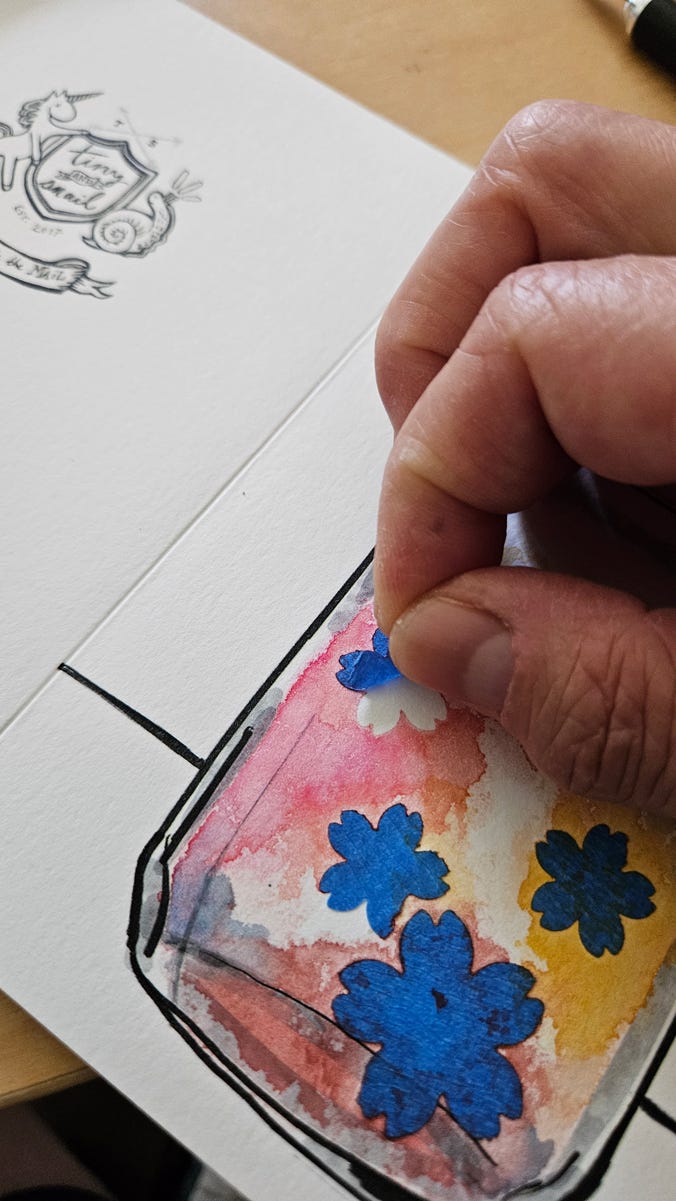 The front of the mason jar card has been painted with blue, yellow and red watercolors over the blue flower-shaped tape masks. A second image shows a detail of fingers peeling off a flower-shaped tape mask.