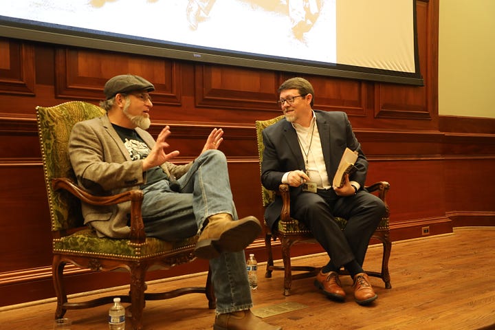 Bob Beatty (left) and Steve Murray (right) sit in chairs. Bob is wearing a hat, a tweed coat with elbow patches, a black Muscle Shoals Sound tshirt, blue jeans, and boots. Steve is in a gray suit with a white shirt. 