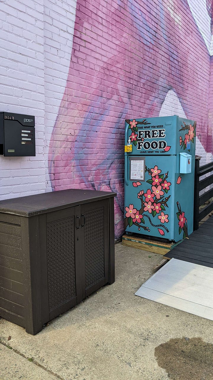 A fridge outside a cement building with lots of colorful flowers painted on it and the words 'FREE FOOD' on it, and a fridge open and full of tons of fresh produce. 