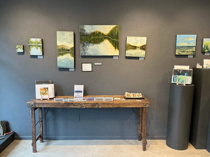 A gallery wall with multiple paintings of a lake reflection