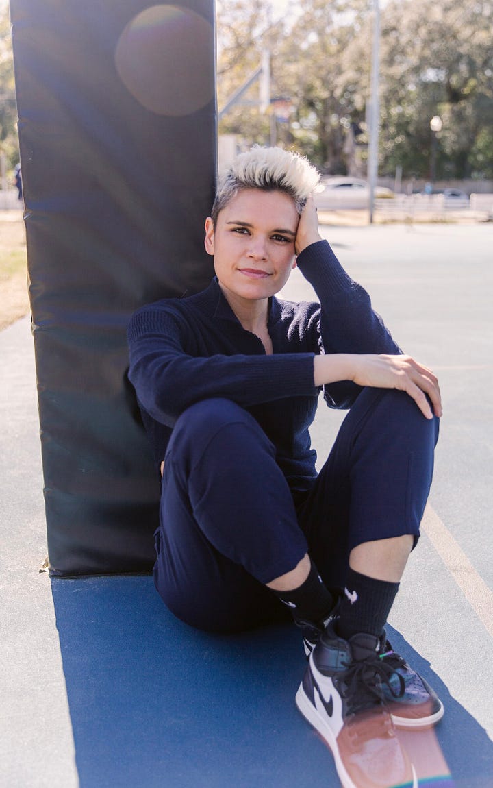 Kate Fagan with short blonde hair sitting casually on the ground. Her book cover for The Three Lives of Cate Kay - red with three shattered images of a woman in a rear view mirror