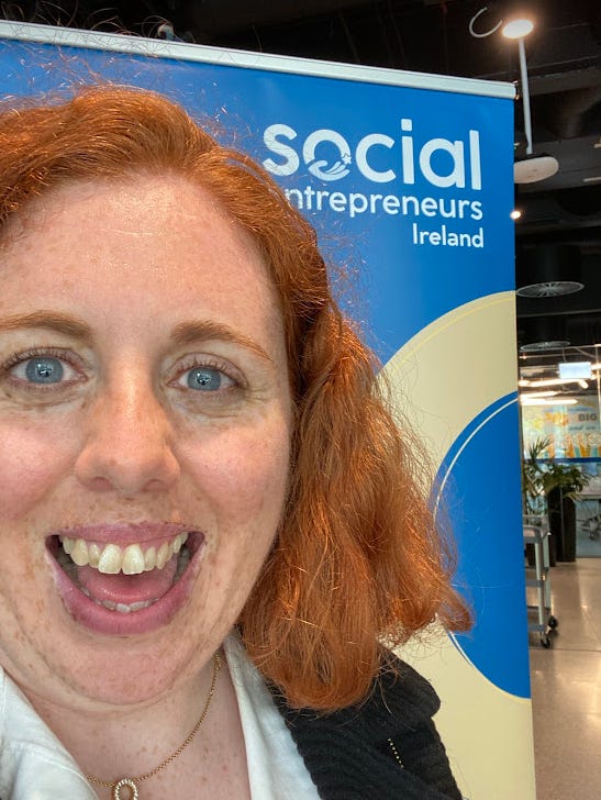 Red-headed woman smiling in front of a Social Entrepreneurs Ireland banner, and a notebook embossed with Social Entrepreneurs Ireland 