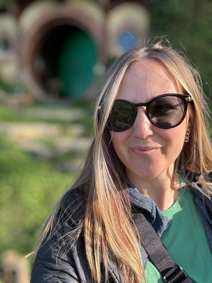 A large garden at the base of Hobbiton with round Hobbit doors tucked into a hillside. A selfie photo of Eija Sumner in front of a blurred background with a green Hobbit door. 