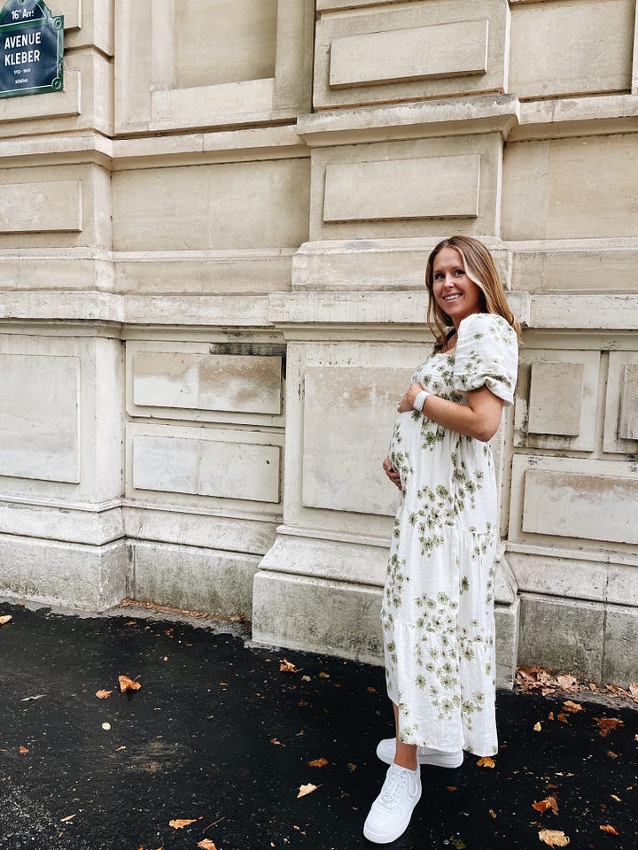 a woman stands in front of an off-white building. she is wearing a cream colored dress with green flowers and white sneakers, and is touching her baby bump and smiling at the camera. 