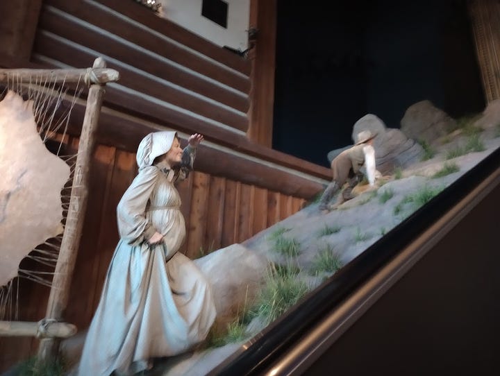 Long elevator with tableau of dry sand and rocks on either side, and a desert view of wagons at top. Indian woman in traditional dress with non-Native man in beard, buckskin, and carrying a rifle, man in wide-brimmed hat clambering up rocky hillside, followed by woman in dress and bonnet. Close view from top of escalator reveals that the passage at top imitates walking through a covered wagonn