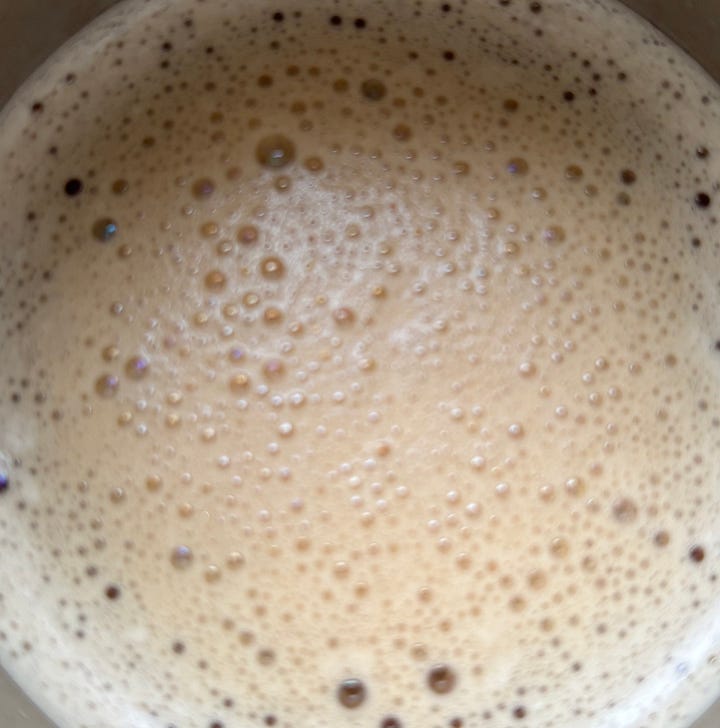 A collage of two images.  On the left, a thermal coffee mug sits in front of a window looking over a snowy scene.  In the right, there's a closeup of the bubble patterns in the coffee milk.