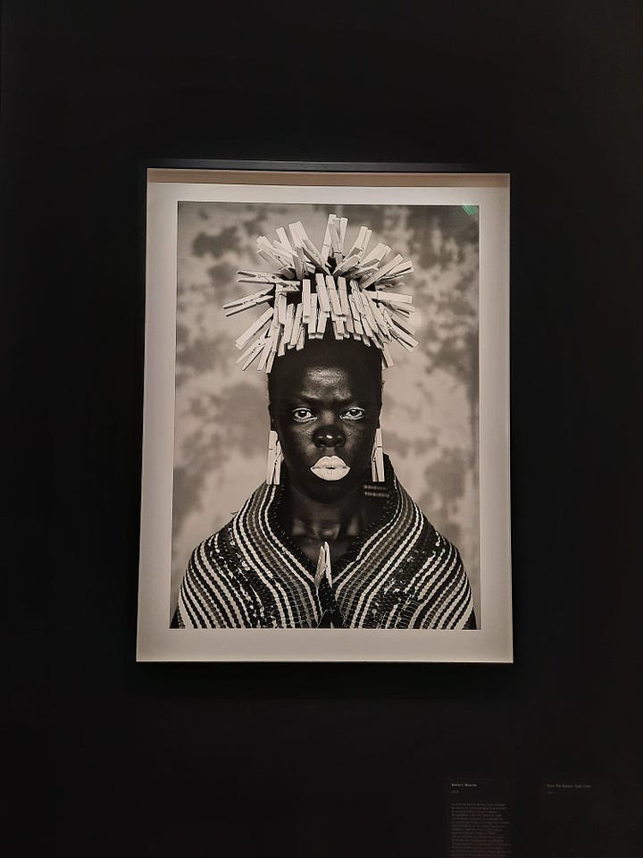 En haut à gauche : Ntozakhe II, Parktown, 2016 © Zanele Muholi. Il s'agit d'un portrait en noir et blanc d'une femme noire dont le regard fixe le haut du cadre. Ses cheveux sont coiffés avec des brosses à cheveux./ En haut à droite :  "LiZa I", 2009 © Zanele Muholi. Il s'agit d'une photographie en noir et blanc d'un couple de femmes s'enlaçant dans des draps, vue de haut, elles sont de dos./ En bas à gauche : vue de l'exposition de l'oeuvre "Faces and Phases" composée de nombreux portraits en noir et blanc./  En bas à droite : "Bester I", 2015 © Zanele Muholi. Il s'agit d'un portrait en noir et blanc d'une femme noire, dont le maquillage laisse la bouche blanche ainsi que les yeux. Ses cheveux sont coiffés de pinces à linge. 