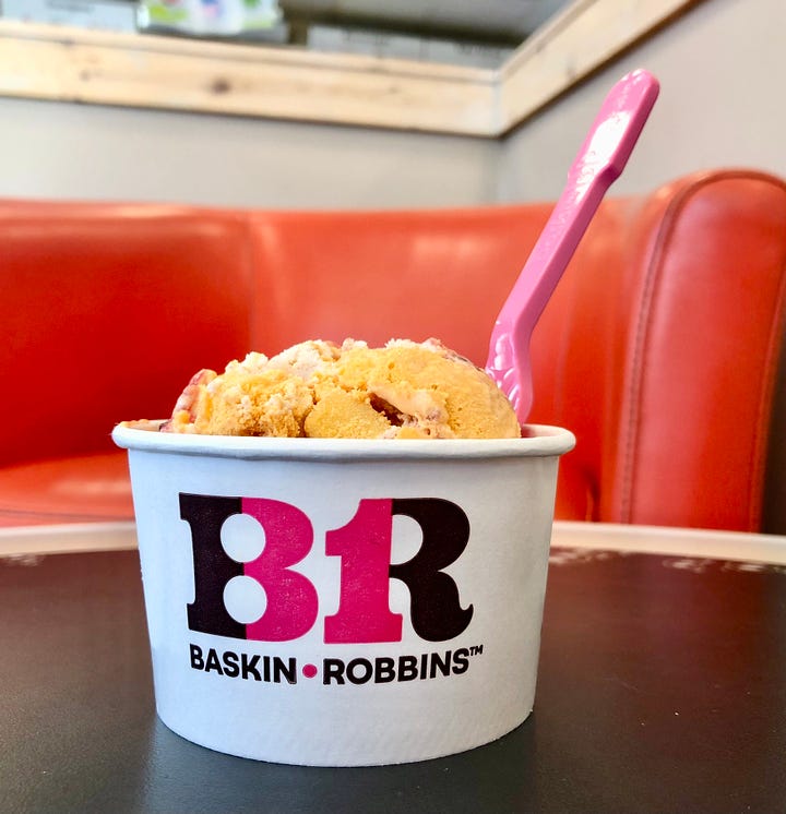 A brightly lit glass case with several different flavors of ice cream in pink and white containers, and a scoop of Turkey Day Fixin's ice cream in a white cup, with the BR/31 logo on front.