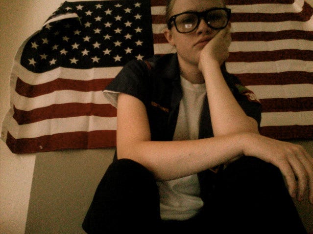 More young soft emo Caley looking disgruntled in front of an American flag or posing in a parking lot with friends.
