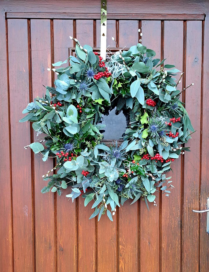 1. Brown door with lush and abundant circular evergreen wreath with red holly berries hanging on it. 2. An image of The Bppo of the Cailleach held infront of a window revealling a white frosty garden in the background.