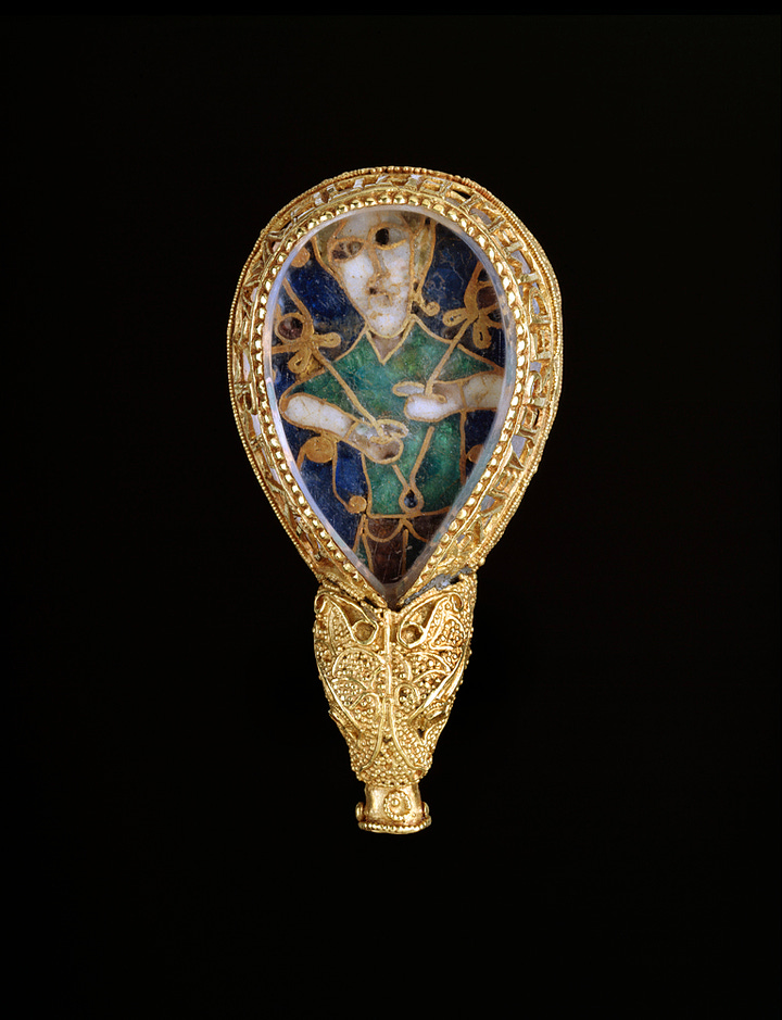 Gold, rock crystal, enamel. Dated to the late 9th century. Gold tear-drop shaped frame holds an enamel base beneath a polished rock crystal, possibly a reused piece from an older object. The separate back plate is held in place by dog-toothed claws, and the jewel terminates in a stylised animal head. The cloisonné enamel inlay depicts a green robed figure holding two plant stems or flowers, and has been interpreted as a personification of sight. The piece may be the head of an aestel or pointer used in the reading of manuscripts. The inscription reads +AELFREDMECH/EH/TGEWYRCAN 'Alfred ordered me to be made'. Description taken from the Woruldhold.
