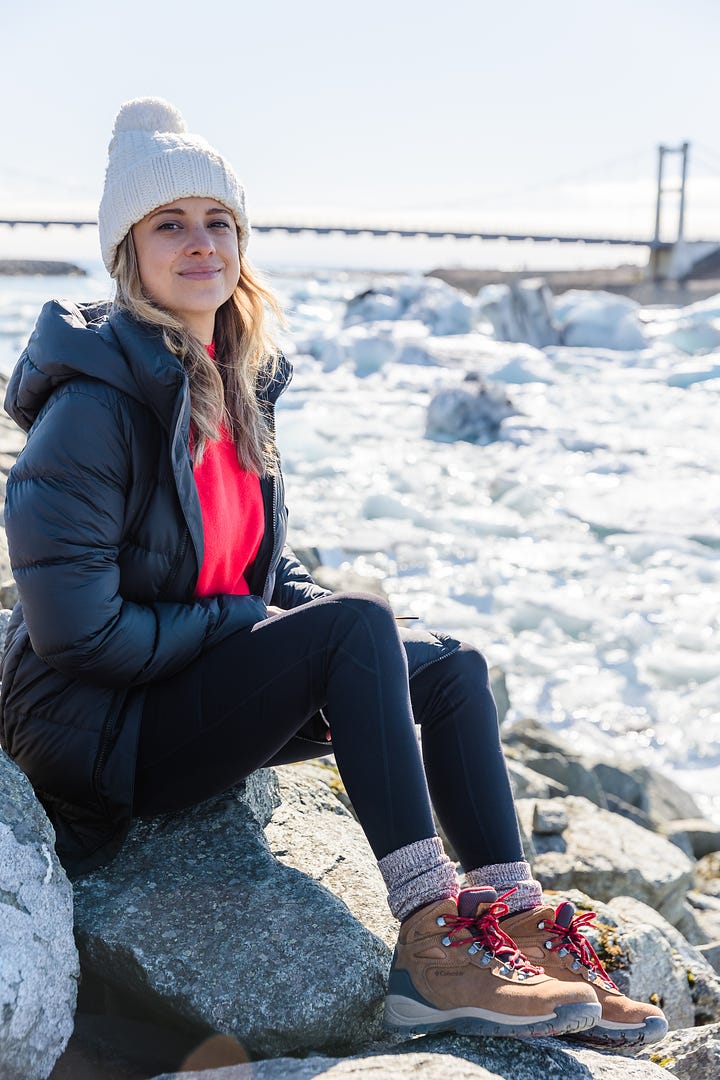 Photo of woman standing along a black sand beach to look at shells and pebbles; woman smiling sitting on a large boulder along a river filled with glacial melt and icebergs with a bridge in the distance; woman laughing and talking at an outdoor picnic table with a group of friends having coffee; selfie photo of two woman smiling wearing sunglasses and winter clothes.