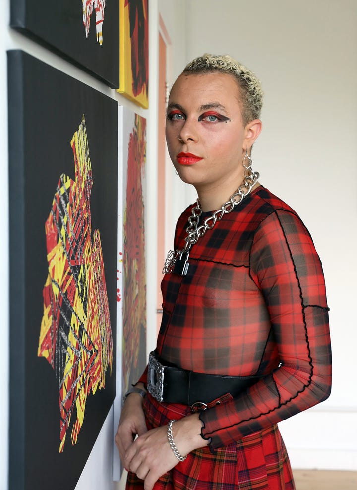 Warren wears a red and black tartan sheer shirt. He had strong red and black eyemakeup, red lipstick and short blonde curly hair. He wears a thick silver padlock chain necklace, a black belt with a silver buckle and stands in front of his artwork
