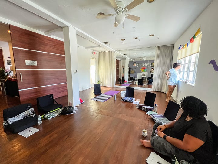 Photo of a lilac painted wall with the word 'synergy' painted on it in white and lotus in all the colors of the rainbow painted on as well; Photo of a wood-floor yoga practice room with low seats on the floor for a group of students, during a break so one student looks out the window and another sittings on the floor; Photo of a mango tree whose leaves are casting light and shadow onto a ochre pathway bordered by potted plants and a string of Tibetan prayer flags hangs overhead; photo of group of 10 women after receiving their Reiki certification, non students and one teacher, some holding up their certificates.